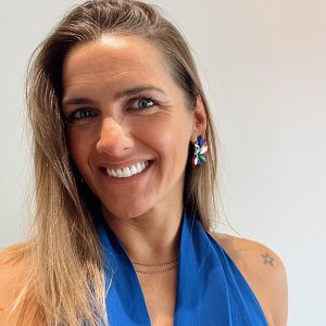 Fotografia de Joana Mineiro - uma mulher de olhos claros e cabelo loiro de sorriso aberto e top azul.O fundo é de uma cor neutra clara, proporcionando um contraste nítido com o indivíduo, o que é comum em retratos profissionais ou corporativos.