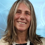 Fotografia de Manuela Francisco - uma mulher de cabelo castanho claro e olhos castanho a sorrir sobre um fundo azul claro. Vestida com uma camisola preta e blazer beje.