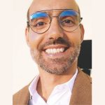 Fotografia de Márcio Martins - um homem sorridente de óculos com olhos castanhos e barba. Vestido com uma camisa branca e um blazer castanho. O fundo é de uma cor neutra clara, proporcionando um contraste nítido com o indivíduo, o que é comum em retratos profissionais ou corporativos.