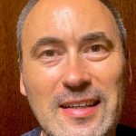 Fotografia de Adelino Couceiro a olhar para a câmra. A pessoa tem olhos castanhos, cabelo e barba grisalha e encontra-se a esboçar um leve sorriso. O fundo é castanho e faz contraste com a cara.