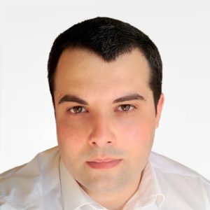 Fotografia do Cláudio Esperança, homem de pele clara, olhos castanhos e cabelo preto, vestido com uma camisa branca. O fundo é de uma cor neutra clara, proporcionando um contraste nítido com o indivíduo, o que é comum em retratos profissionais ou corporativos.