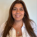 Fotografia de Renata Botelho, mulher de tonalidade morena com cabelo comprido castanho e olhos castanhos a esboçar um sorriso para a câmara. Encontra-se vestida com uma camisa bordada bordada e top beje e possui um colar ao pescoço. O fundo é de uma cor neutra clara, proporcionando um contraste nítido com o indivíduo.