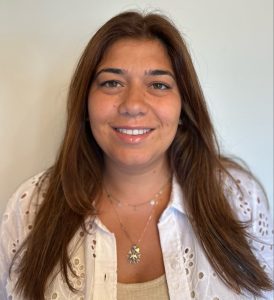 Fotografia de Renata Botelho, mulher de tonalidade morena com cabelo comprido castanho e olhos castanhos a esboçar um sorriso para a câmara. Encontra-se vestida com uma camisa bordada bordada e top beje e possui um colar ao pescoço. O fundo é de uma cor neutra clara, proporcionando um contraste nítido com o indivíduo.