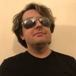 Fotografia de João de Sousa e Silva, homem com óculos escuros e t-shirt preta. O fundo é de uma cor amarela clara, proporcionando contraste com o indivíduo, o que é comum em retratos profissionais ou corporativos.