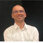 Fotografia de Pedro Alves com camisa branca. A pessoa tem cabelo muito curto e relativamente grisalho, está barbeada, a sorrir e usa óculos. O fundo é totalmente negro, aparentando ser um palco de um evento e proporcionando um contraste totalmente nítido com o indivíduo.