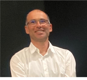 Fotografia de Pedro Alves com camisa branca. A pessoa tem cabelo muito curto e relativamente grisalho, está barbeada, a sorrir e usa óculos. O fundo é totalmente negro, aparentando ser um palco de um evento e proporcionando um contraste totalmente nítido com o indivíduo.