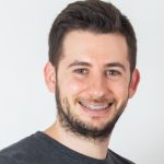 Fotografia de Rafael Coelho, homem com cabelo escuro e olhos castanhos de sorriso aberto e barba. Vestido com uma t-shirt cinzenta. O fundo é de uma cor neutra clara, proporcionando um contraste nítido com o indivíduo, o que é comum em retratos profissionais ou corporativos.