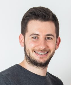 Fotografia de Rafael Coelho, homem com cabelo escuro e olhos castanhos de sorriso aberto e barba. Vestido com uma t-shirt cinzenta. O fundo é de uma cor neutra clara, proporcionando um contraste nítido com o indivíduo, o que é comum em retratos profissionais ou corporativos.