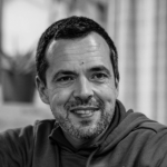 Fotografia de António Almeida - um homem de cabelo escuro e barba grisalha cuidada a sorrir levemente, com um ar descontraído. A fotografia é a preto e branco.