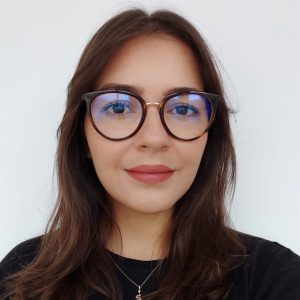 Fotografia de Daniele Miranda, uma mulher de cabelo castanho e olhos castanhos, com um sorriso suave e expressão acolhedora. Usa óculos de aros arrendondados e uma camisola preta, conferindo um ar profissional e moderno.