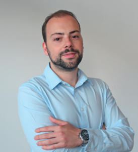 Fotografia de Hugo Peixe, homem com cabelo e barba castanho escuro. O fundo é de uma cor neutra clara, proporcionando um contraste nítido com o indíviduo, o que é comum em retratos profissionais ou corporativos.