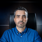 Fotografia de Jorge Guerreiro - um homem de expressão séria e olhar direto, de cabelo curto preto e barba aparada. Usa uma camisa azul transmitindo um ar formal.