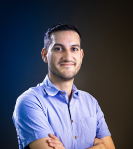 Fotografia de Marcelo Gonçalves, um homem de cabelo curto preto e olhos castanhos escuros. Apresenta barba ligeira, braços cruzados e encontra-se a sorrir levemente para a câmara. O fundo apresenta um degradé escuro, entre o azul e o castanho, o que contribui para destacar a figura principal com um toque moderno e profissional.