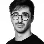 Fotografia de Miguel Silva - um homem de cabelo escuro, com expressão neutra e ligeiramente curiosa. Usa óculos redondos de armação escura e exibe uma barba curta e cuidada. A fotografia é a preto e branco.