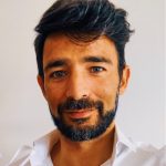 Fotografia de Bruno Godinho - homem de cabelo preto com barba bem cuidada com expressão serena e a sorrir ligeiramente. Veste uma camisa branca, conferindo um aspeto profissional.