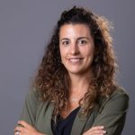 Fotografia de Filipa Sena Nunes, uma mulher de cabelo castanho encaracolado e olhos castanhos a sorrir para a câmara. Usa um casaco verde e camisola preto em contraste com o fundo cinza, comum em fotos corporativas.