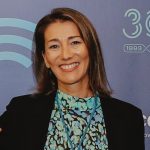 Fotografia de Isabel Varajão, uma mulher de cabelo castanho e olhos castanhos. Apresenta um largo sorriso para a câmara e veste um casaco preto sobre uma camisola com padrões verdes, conferindo um ar moderno e corporativo.