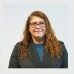 Fotografia de Namar Faria, uma mulher de cabelo castanho claro encaracolado, com um sorriso suave e expressão acolhedora. Usa óculos de aros vermelhos e um casaco preto sobre uma camisola azul, conferindo um ar profissional e moderno.