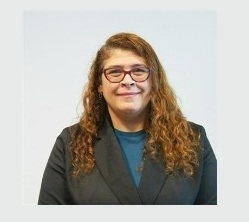 Fotografia de Namar Faria, uma mulher de cabelo castanho claro encaracolado, com um sorriso suave e expressão acolhedora. Usa óculos de aros vermelhos e um casaco preto sobre uma camisola azul, conferindo um ar profissional e moderno.