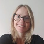 Fotografia de Mafalda Goulart - uma mulher de cabelo loiro e olhos azuis sorridente para a câmera. Usa óculos de armação redonda e o fundo é de cor neutra, típica em fotos corporativas.