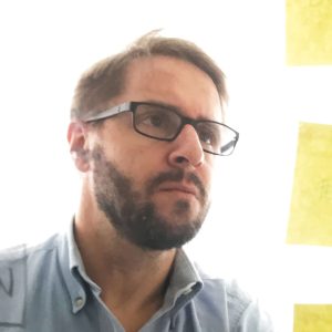 Fotografia de Rogério Albertino - um homem de cabelo escuro e barba cuidada, com expressão neutra. Usa óculos rectangulares de armação escura e encontra-se de perfil para a foto.