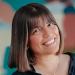 Fotografia de Sara Sirvoicar - uma mulher de cabelo castanho e olhos castanhos sorridente para a câmara, Usa franja e encontra-se com a cabeça levemente inclinada.