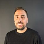 Fotografia de João Brito, um homem de cabelo castanho e olhos castanhos a esboçar um sorriso para a câmera. Veste uma camisola preta e o fundo é cinzento, comum em retratos corporativos.