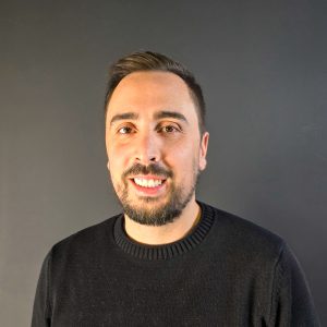 Fotografia de João Brito, um homem de cabelo castanho e olhos castanhos a esboçar um sorriso para a câmera. Veste uma camisola preta e o fundo é cinzento, comum em retratos corporativos.