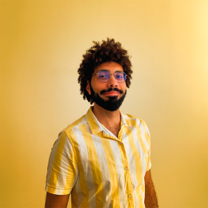 Fotografia de Gabriel Patrocinio - um homem cabelo castanho e olhos castanhos. Tem barba cuidada e encontra-se a sorrir para a câmara, Usa uma camisa às riscas amarela e branca que combina com o fundo igualmente amarelo.