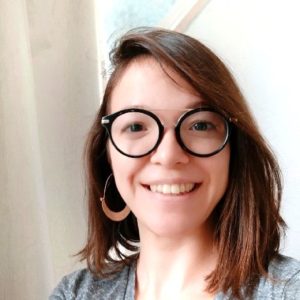 Fotografia de Natali Nascimento, uma mulher de olhos e cabelo castanho. Usa óculos de armação preta de formato redondo e encontra-se a sorrir para a foto.