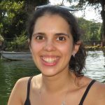 Fotografia de Sandra Costa, uma mulher de olhos castanho e com o cabelo castanho apanhado. Esboça um largo sorriso para foto e o fundo é em ambiente outdoor.