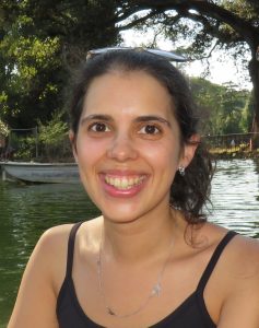 Fotografia de Sandra Costa, uma mulher de olhos castanho e com o cabelo castanho apanhado. Esboça um largo sorriso para foto e o fundo é em ambiente outdoor.