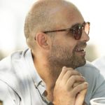 Fotografia de Gonçalo Maria, um homem de perfil com óculos de sol a esboçar um sorriso. Veste uma camisa cinzenta clara e apresenta um ar descontraído.