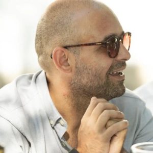 Fotografia de Gonçalo Maria, um homem de perfil com óculos de sol a esboçar um sorriso. Veste uma camisa cinzenta clara e apresenta um ar descontraído.