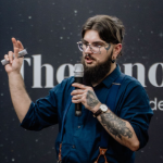 Fotografia de Pedro Pereira, um homem de cabelo e barba castanha, com óculos. Veste uma camisa azul e apresenta diversas tatuagens e piercings. A foto é tirada num contexto de palestra, conferindo um ar profissional.