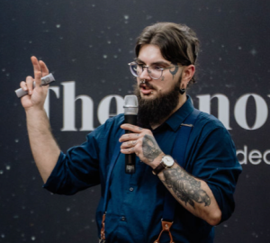 Fotografia de Pedro Pereira, um homem de cabelo e barba castanha, com óculos. Veste uma camisa azul e apresenta diversas tatuagens e piercings. A foto é tirada num contexto de palestra, conferindo um ar profissional.