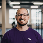 Fotografia de Alexandre Santos, hum homem com cabelo e olhos castanho e barba cuidada a sorrir para a câmara. Veste uma camisola azul e usa óculos de armação preta rectangular.