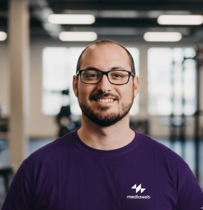 Fotografia de Alexandre Santos, hum homem com cabelo e olhos castanho e barba cuidada a sorrir para a câmara. Veste uma camisola azul e usa óculos de armação preta rectangular.