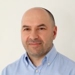 Fotografia de Ricardo Alves, homem de olhos castanhos e barba aparada com expressão serena e a sorrir ligeiramente. Veste uma camisa azul e o fundo é branco, conferindo um aspeto profissional comum em fotos corporativas.