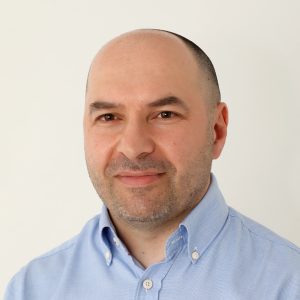 Fotografia de Ricardo Alves, homem de olhos castanhos e barba aparada com expressão serena e a sorrir ligeiramente. Veste uma camisa azul e o fundo é branco, conferindo um aspeto profissional comum em fotos corporativas.