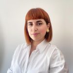 Fotografia de Joana Duarte, uma mulher de cabelo ruivo e olhos castanhos a sorrir levemente para a foto. Veste uma camisa branca e o fundo é neutro, comum em fotos corporativas.