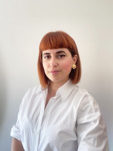 Fotografia de Joana Duarte, uma mulher de cabelo ruivo e olhos castanhos a sorrir levemente para a foto. Veste uma camisa branca e o fundo é neutro, comum em fotos corporativas.