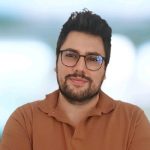 Fotografia de Rui Joel Costa, um homem de cabelo e olhos castanhos escuros. Usa óculos de aros arredondados e veste uma camisola castanha.