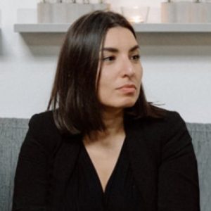 Fotografia de Carolina Alves, uma mulher de olhos e cabelo castanho escuro. Veste uma camisola preta e encontra-se de perfil para a câmara.