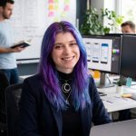 Fotografia de Jéssica Facchin, uma mulher de olhos castanhos e cabelo roxo com um sorriso largo para a câmera.